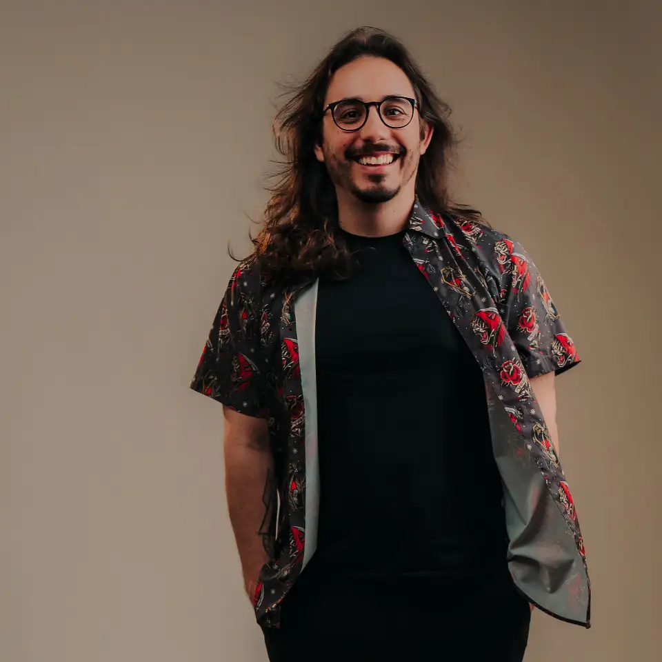 Headshot portrait of João Dias seated on a brown leather armchair. He has long wavy hair, glasses, a mustache, and visible forearm tattoos, wearing a short-sleeved black polo shirt against a warm, neutral backdrop.