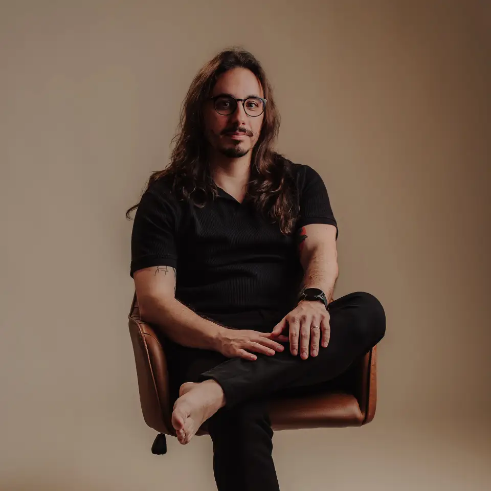 Headshot portrait of João Dias seated on a brown leather armchair. He has long wavy hair, glasses, a mustache, and visible forearm tattoos, wearing a short-sleeved black polo shirt against a warm, neutral backdrop.